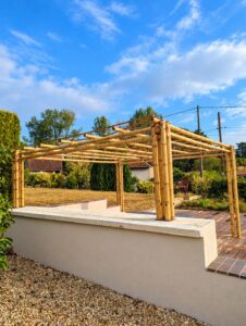 Pergola en bambou autoportée installée sur une terrasse, avec poteaux multiples, charpente ouverte et jardin paysager en arrière-plan.