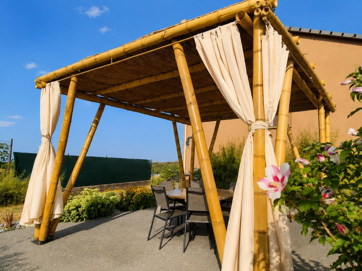 Pergola en bambou avec couverture naturelle et rideaux, aménagée sur une terrasse de jardin avec un espace repas.