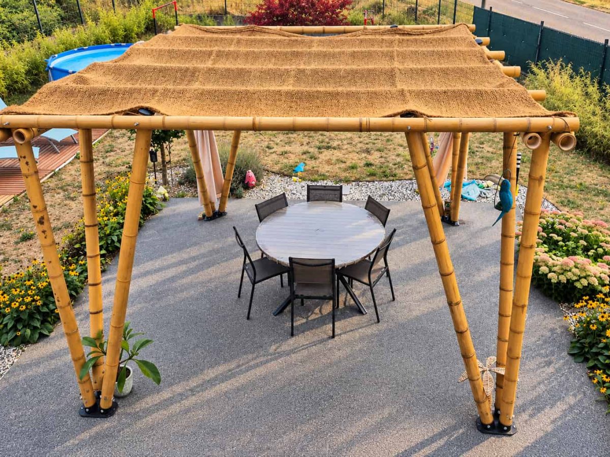 Pergola en bambou autoportée installée sur une terrasse avec table et chaises, entourée de plantations et de massifs fleuris.