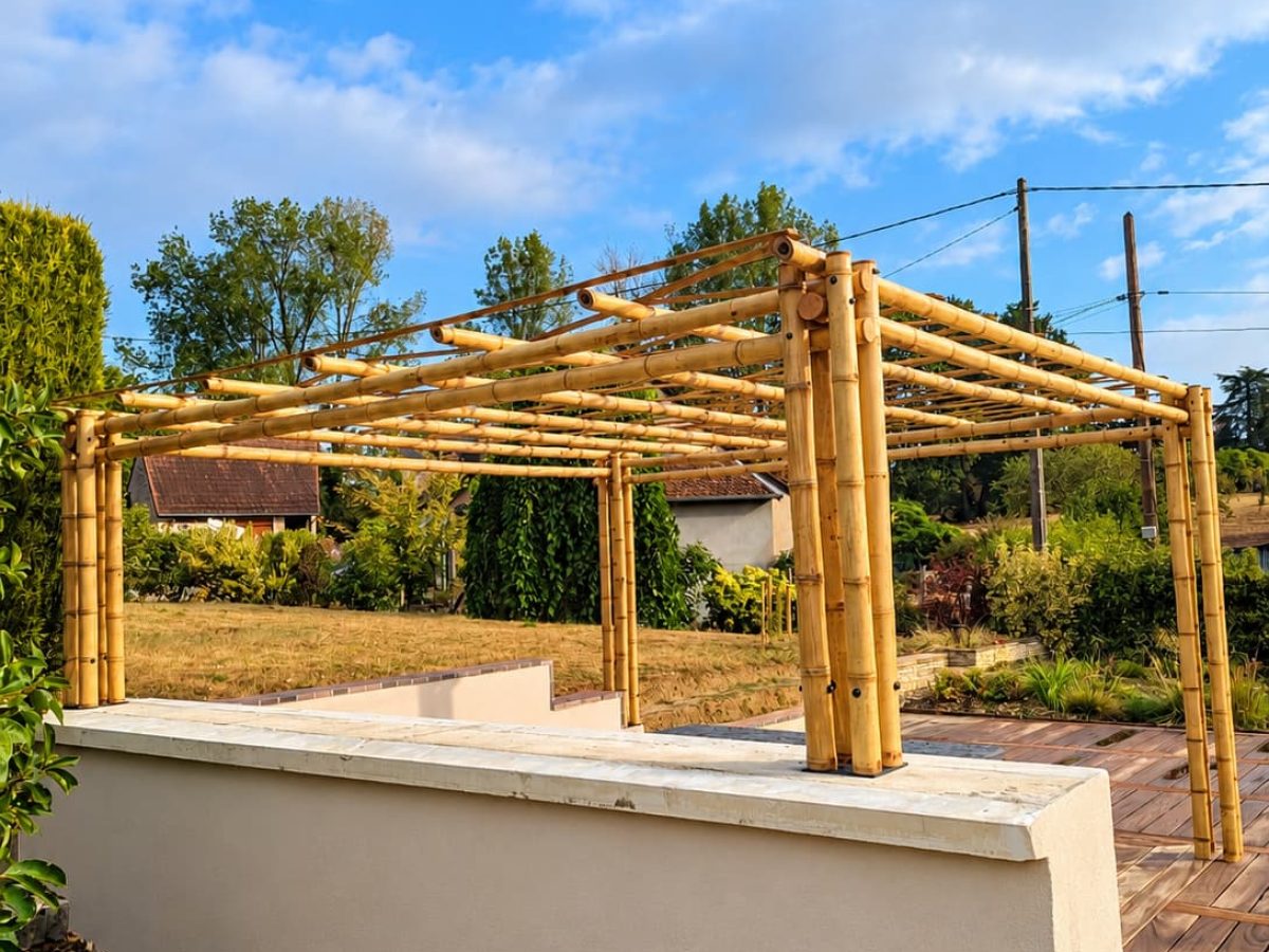 Pergola en bambou autoportée installée sur une terrasse, avec poteaux multiples, charpente ouverte et jardin paysager en arrière-plan.