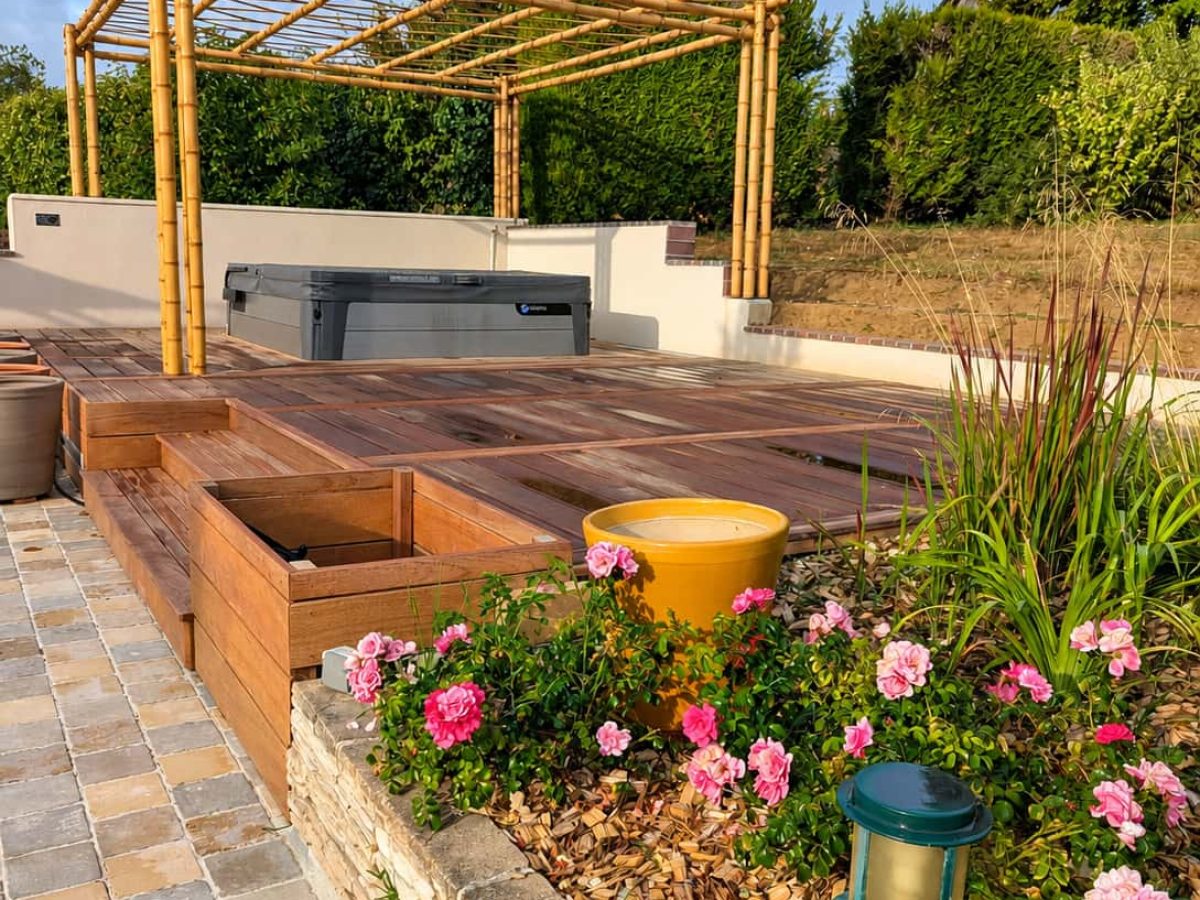 Pergola en bambou installée sur une terrasse en bois avec spa, entourée de massifs fleuris et de végétation.