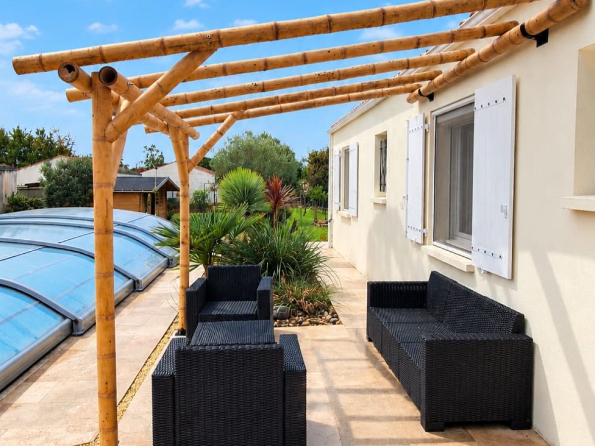 Pergola en bambou adossée à une maison avec terrasse aménagée, salon de jardin, végétation et piscine.